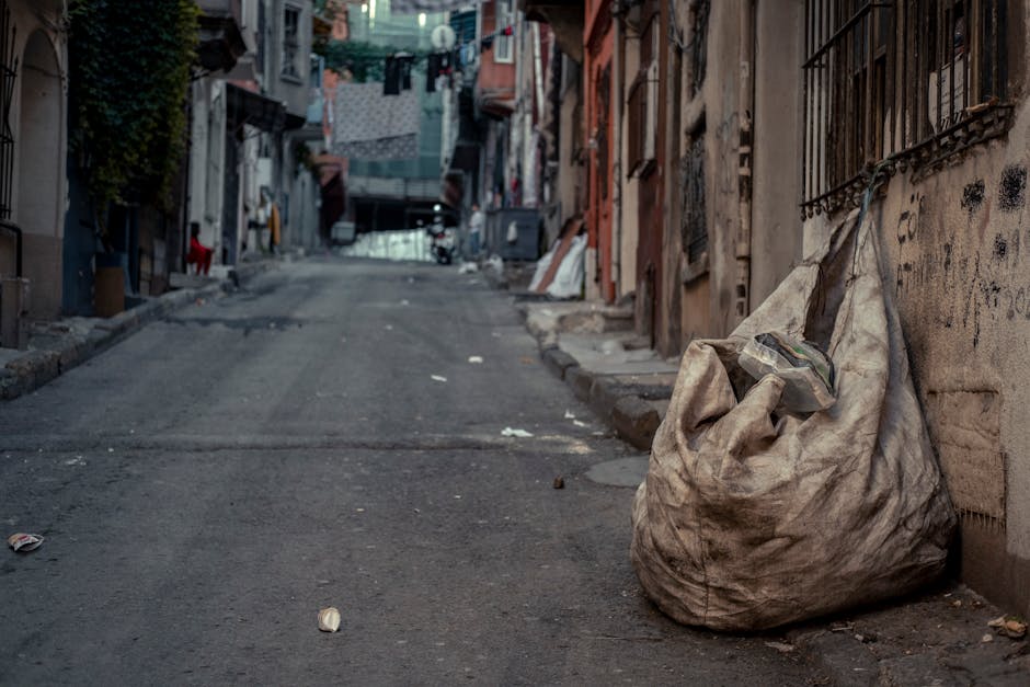 A large, weathered, off-white plastic trash bag rests on a narrow street pavement, leaning against a wall with graffiti and iron window bars. The bag appears full and slightly bulging, with its top gathered and tied. Surrounding the bag, there is some scattered litter, including small pieces of paper and debris. The street surface is rough, uneven, and darkened with dirt, showing signs of wear and patchwork repairs. In the background, the street extends uphill, lined with tightly packed residential buildings with various textures such as brick, plaster, and metal. Some buildings have laundry hanging overhead on clotheslines, and others display windows with shutters or bars. The scene is dimly lit, possibly during late afternoon or early evening, with a somewhat gritty urban atmosphere characteristic of densely populated areas where private waste collection services like House Clearance Camden are often employed for managing rubbish in tight, narrow streets. The overall setting emphasizes the importance of proper waste handling in urban environments with limited space for rubbish accumulation.
