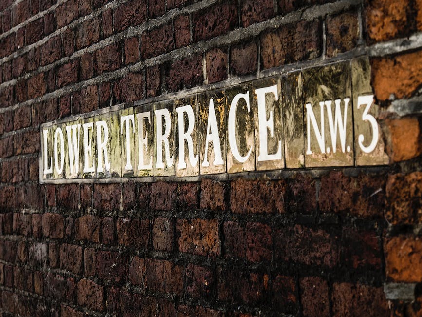 A close-up view of a vintage street sign mounted on a weathered red brick wall, reading 'LOWER TERRACE NW3'. The sign is composed of individual ceramic tiles with a glossy finish, featuring white embossed lettering against a darker background, with some tiles showing signs of moss or dirt accumulation. The brickwork surrounding the sign is uneven, with reddish-brown and dark tones, displaying typical texture of aged masonry with visible mortar joints. The lighting is natural, illuminating the sign and highlighting the rustic surface of the bricks. The scene suggests an urban setting, possibly within a residential or historical area, where signage indicates the street name and postal district. This environment may relate to private property or on-site street signage, relevant for contexts involving property clearance or local area identification, such as those managed by companies like House Clearance Camden, offering alternative waste handling or rubbish removal services in such neighbourhoods.