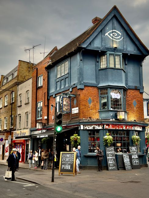 A three-storey corner building with a distinctive blue-painted upper level and brickwork on the lower sections stands at a busy street intersection. The upper part features a gabled roof with dark tiles and large bay windows beneath the eaves, complemented by a white eye symbol painted on the façade. The ground floor houses a pub with outdoor chalkboard signs advertising drink specials, surrounded by potted plants on windowsills and storefront displays. Several pedestrians are gathered outside, some waiting to enter, while others walk past on the pavement. A traffic light showing green is positioned in front of the building, with other shops and residential buildings visible in the background. The overall scene is under a cloudy sky, with natural daylight illuminating the street. The setting reflects an urban environment where independent venues and local businesses, such as House Clearance Camden, may facilitate alternative waste handling or on-site clearance services to complement conventional rubbish removal options.