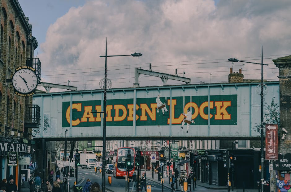 A busy street scene in Camden during daytime features a large, rectangular railway bridge with a weathered green and orange sign displaying 'CAMDEN LOCK' in bold letters in the foreground. The bridge is constructed from metal panels with visible rivets and a slightly rusted finish, supported by brick columns on either side. Below the bridge, there is a mix of pedestrians, some waiting at a crosswalk, and vehicles such as a red double-decker bus and cars moving along the street, indicating typical urban activity. Surrounding the scene are various street furniture elements, including traffic lights, street lamps, and directional signage, with some graffiti visible on nearby walls. On the left side of the image, a large antique-style clock is mounted on a building, showing the time as approximately 11:10. The sky above is partly cloudy, with patches of light filtering through the clouds, adding a diffused lighting to the scene. The environment appears vibrant and dynamic, with a diverse crowd, reflecting the cultural and commercial character of Camden. This visual depiction relates to private waste handling and on-site clearance needs common in bustling urban areas like Camden, with House Clearance Camden possibly involved in rubbish removal services for commercial or residential properties in such locations.