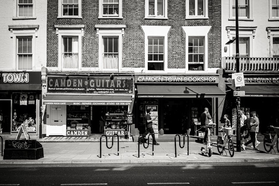 A black-and-white street-level photograph of a row of commercial storefronts on a busy urban pavement, featuring a music store named 'Camden Guitars' with large signage displaying 'Music - Repairs - Exchange - Studio,' and a second shop labeled 'Camden Town Bookshop' with a covered entrance. In front of the shops, several pedestrians are walking along the sidewalk, some carrying shopping bags or backpacks, and one person appears to be looking at their phone. The street includes a bicycle rack with multiple bicycles parked, and a streetlamp stands on the right side, casting a shadow on the pavement. The building facades are made of brick with large windows on the upper floors, and the ground floors have awnings and signage indicating retail activity. The scene is typical of an active commercial area, where external independent collection or disposal of waste or rubbish is likely relevant, as part of everyday waste management practices managed by local or private service providers like House Clearance Camden.