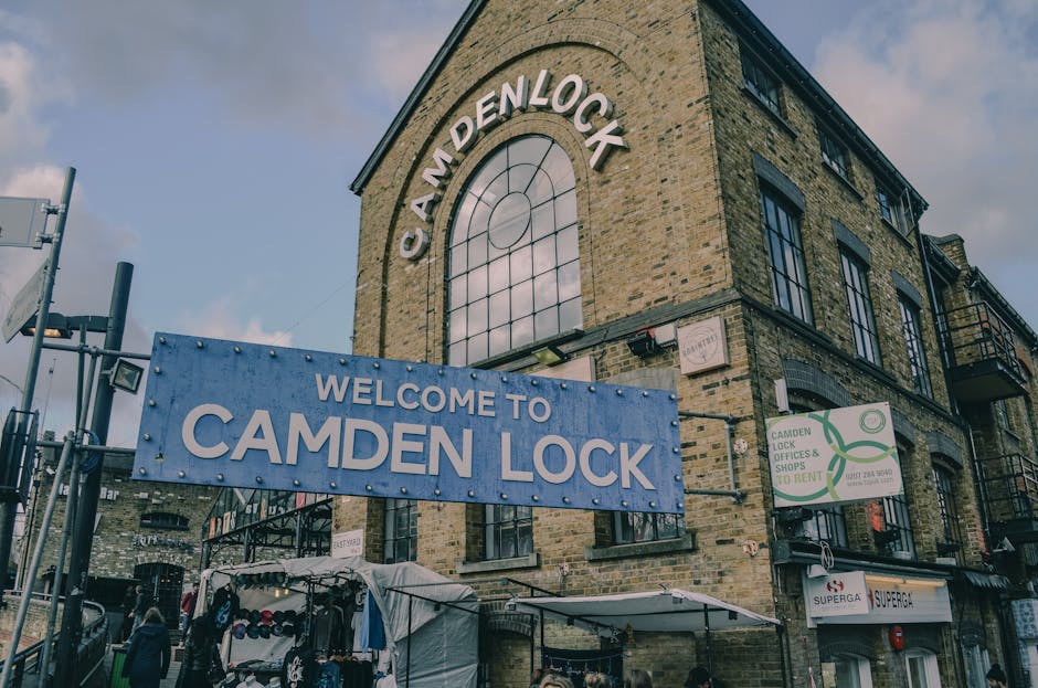 The image shows a multi-story brick building with large, dark metal-framed windows, located on a city street. The building features a prominent arched window with a metal grid design on the upper floor, above a wide blue sign that reads 'WELCOME TO CAMDEN LOCK' in white capital letters. To the right of the main entrance, there is a smaller sign indicating Camden Lock's shops and rental offices, with green accent graphics. In the foreground, a market stall with a white canopy displays an array of clothing and merchandise, supported by metal poles. Adjacent to the stall, a few pedestrians can be seen walking along the sidewalk. The scene is outdoors and illuminated by natural daylight, with a partly cloudy sky overhead. The overall setting suggests a lively, urban environment typical of a popular market or shopping area, where independent collections or private waste disposal methods might be employed when managing rubbish from retail activities. House Clearance Camden often provides rubbish removal services in such busy commercial and residential settings, supporting alternative waste handling solutions in Camden.
